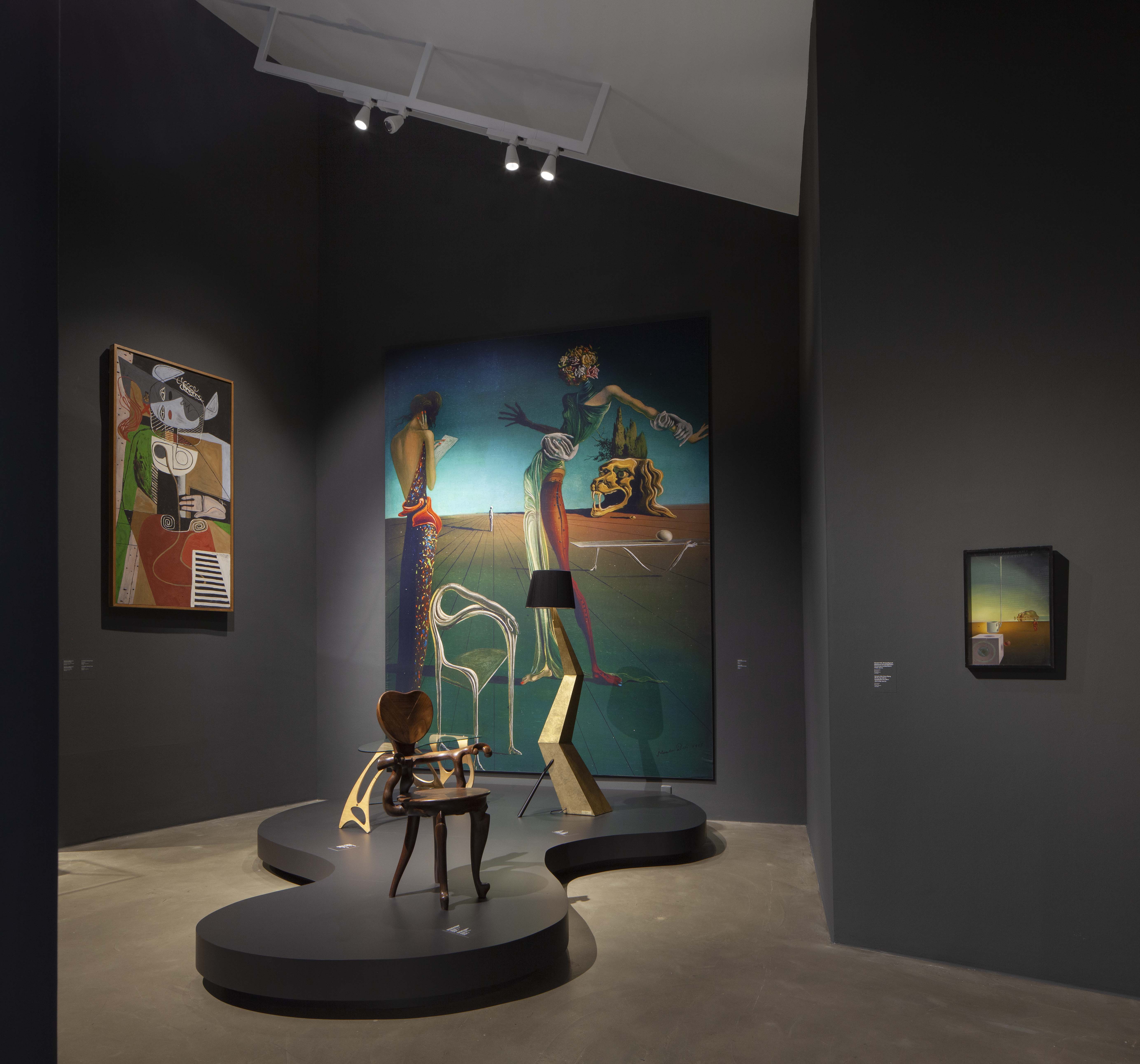Blick in die Ausstellung. Links: Le Corbusier, Taureau VI (1954), an der Rückwand das Bild «Femme à tête des roses» (1935) und rechts «Riesige fliegende Mokkatasse mit unerklärlicher Fortsetzung von fünf Metern Länge» (1944/45), beide von Salvador Dalí. Auf dem Sockel hinten der Tisch Arabesco von Carlo Mollino (1950), die Stehleuchte  aus vergoldetem Holz «Bracelli» (Salvador Dalí, 1937) und vorne der Armlehnstuhl aus Holz für die Casa Calvet von Antoni Gaudí, 1900/1901.