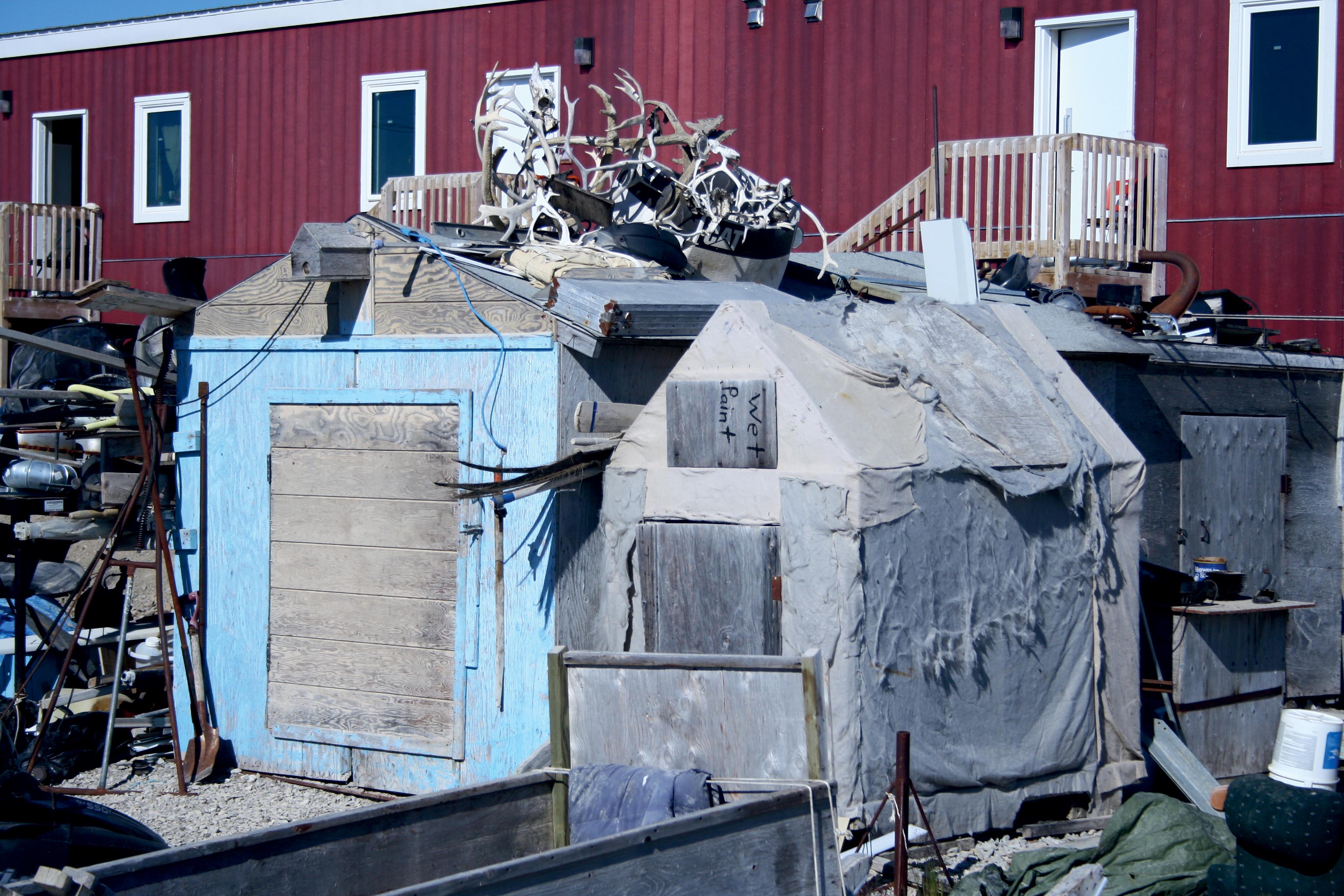 Standard et non-standard : cabanes pour le stockage du matériel de chasse et de pêche avec, en arrière-plan, le logement social proposé par le gouvernement canadien, village d’Igloolik