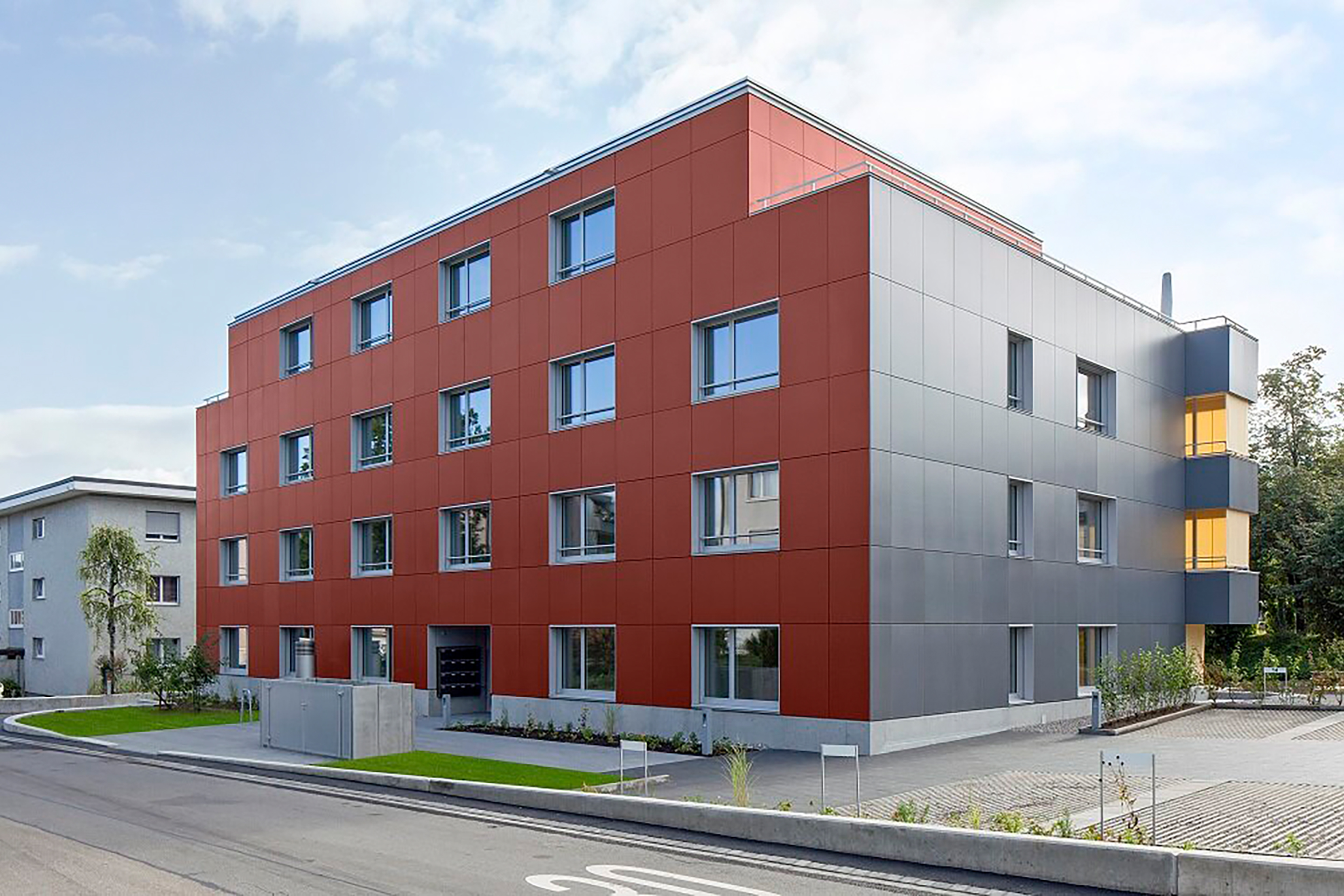 L’edificio abitativo progettato da Viriden+Partners in posizione centrale ad Affoltern am Albis è un volume di tre piani con soffitta. A destra un dettaglio della facciata fotovoltaica di colore rosso e grigio.