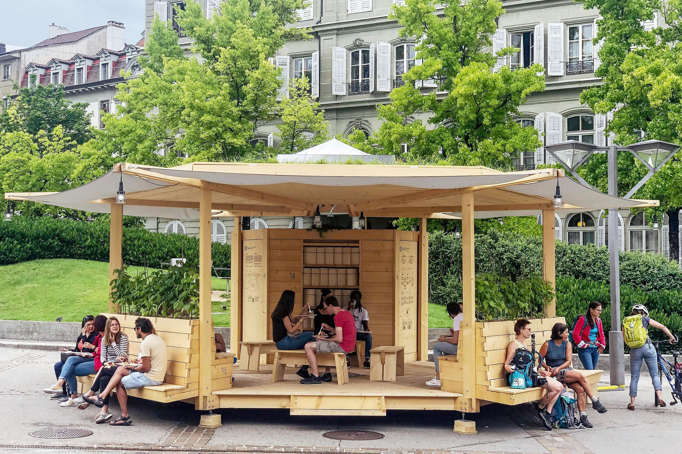 Le pavillon installé en juillet 2021 sur la place Python à Fribourg. Diamètre de la toiture avec le prolongement des toiles d’ombrage: 10 m, hauteur entre le plancher et la toiture: 2,5 m, hauteur maximale : 3,8 m. Sa structure modulaire permet de le démonter, le transporter puis le remonter sur un nouvel emplacement en ~1,5 jours.