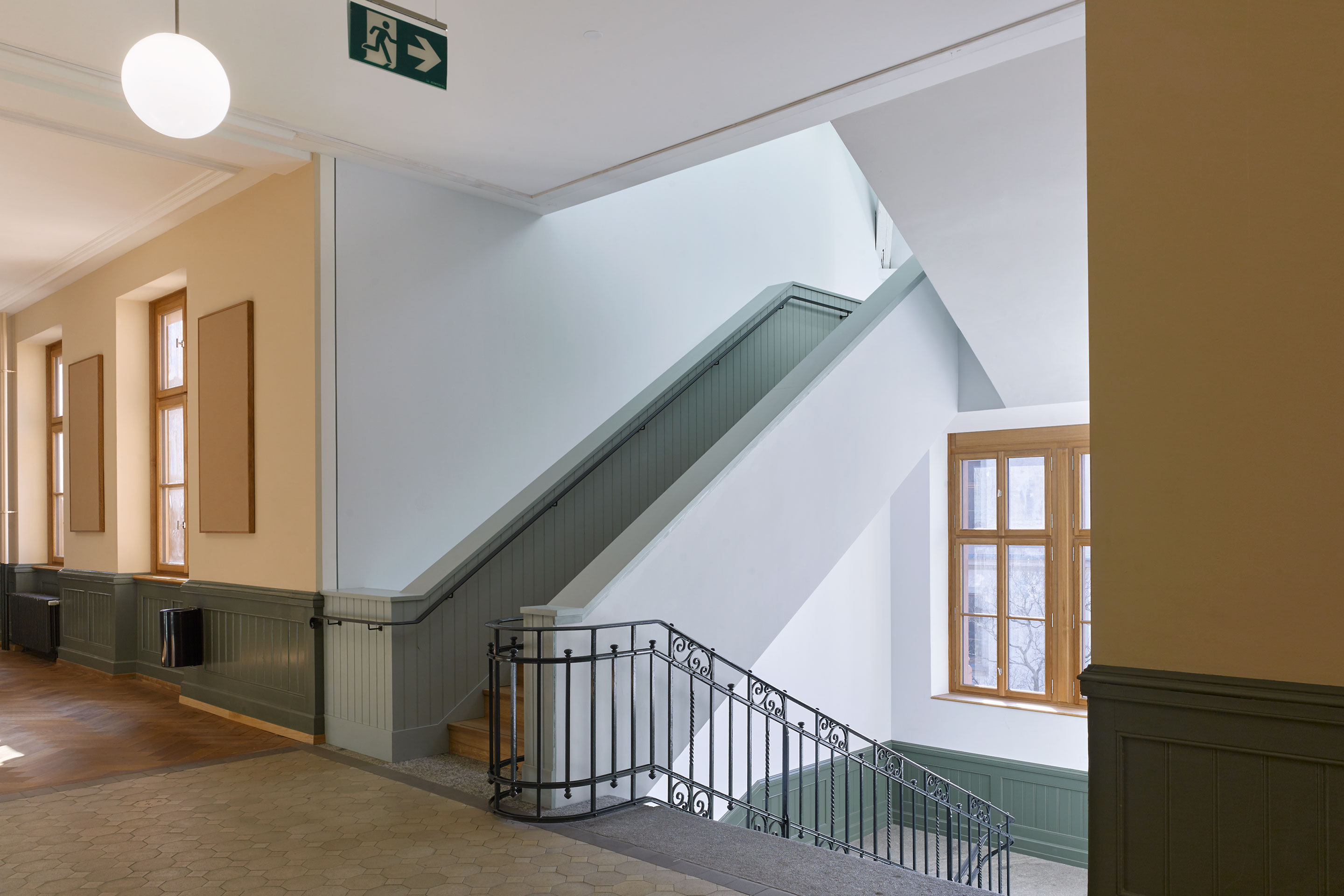 Der neue Übergang ins Dachgeschoss. Aus dem bestehenden repräsentativen Treppenhaus mit seinem kunsteisernen Handlauf entwickeln sich zwei schmalere Betonläufe mit frei kragendem Podest. Die massiven Brüstungen machen den Wechsel vom Bestand zum neuen Element spürbar, gleichzeitig ist durch das Anknüpfen an das bestehende Treppenhaus die Weiterführung selbstverständlich.