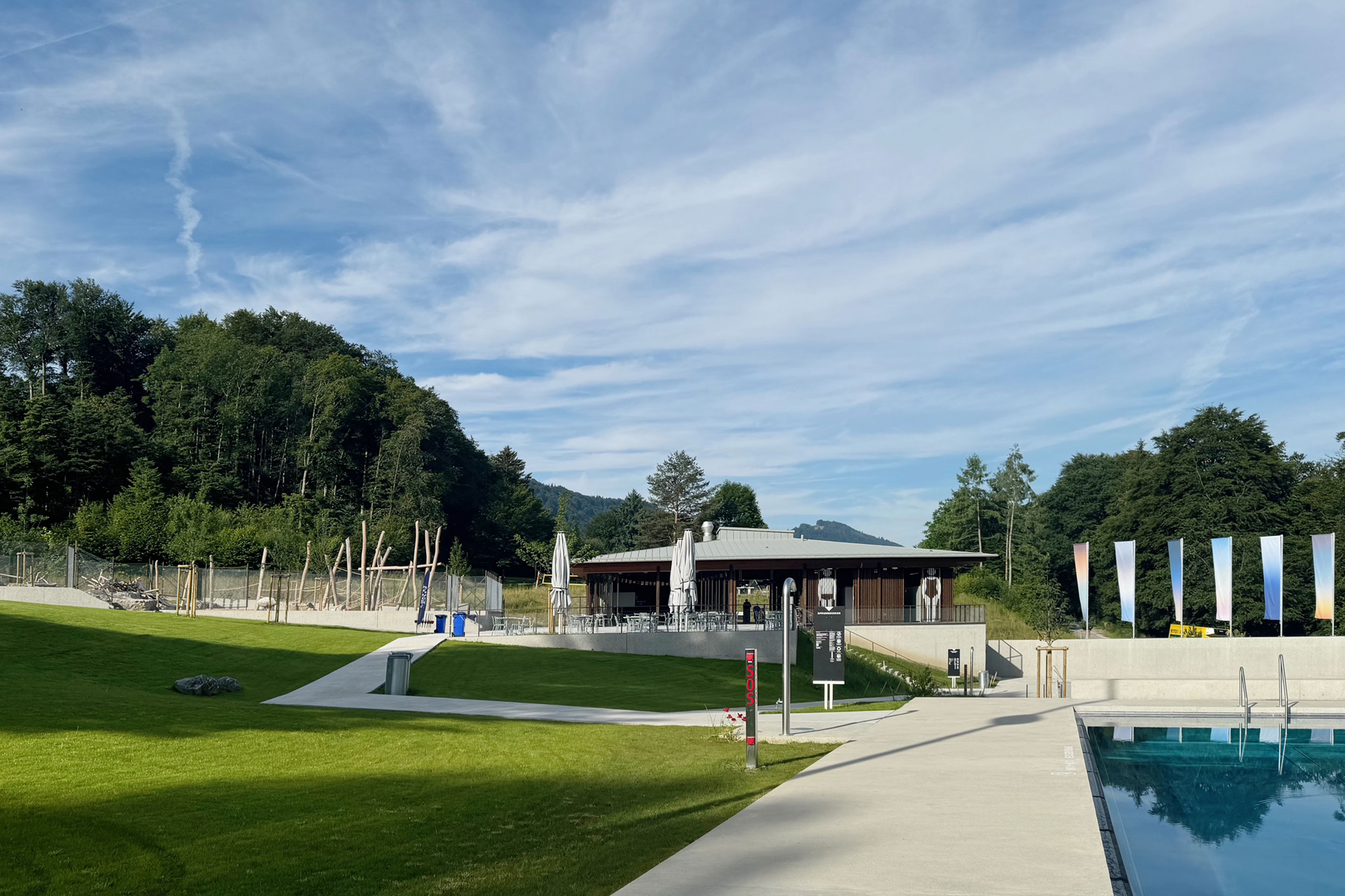 Blick vom Sprungbecken zum Eingangsgebäude. Dieses wird flankiert vom Spielplatz (links) und den Flaggen des Kunst-und-Bau-Objekts.