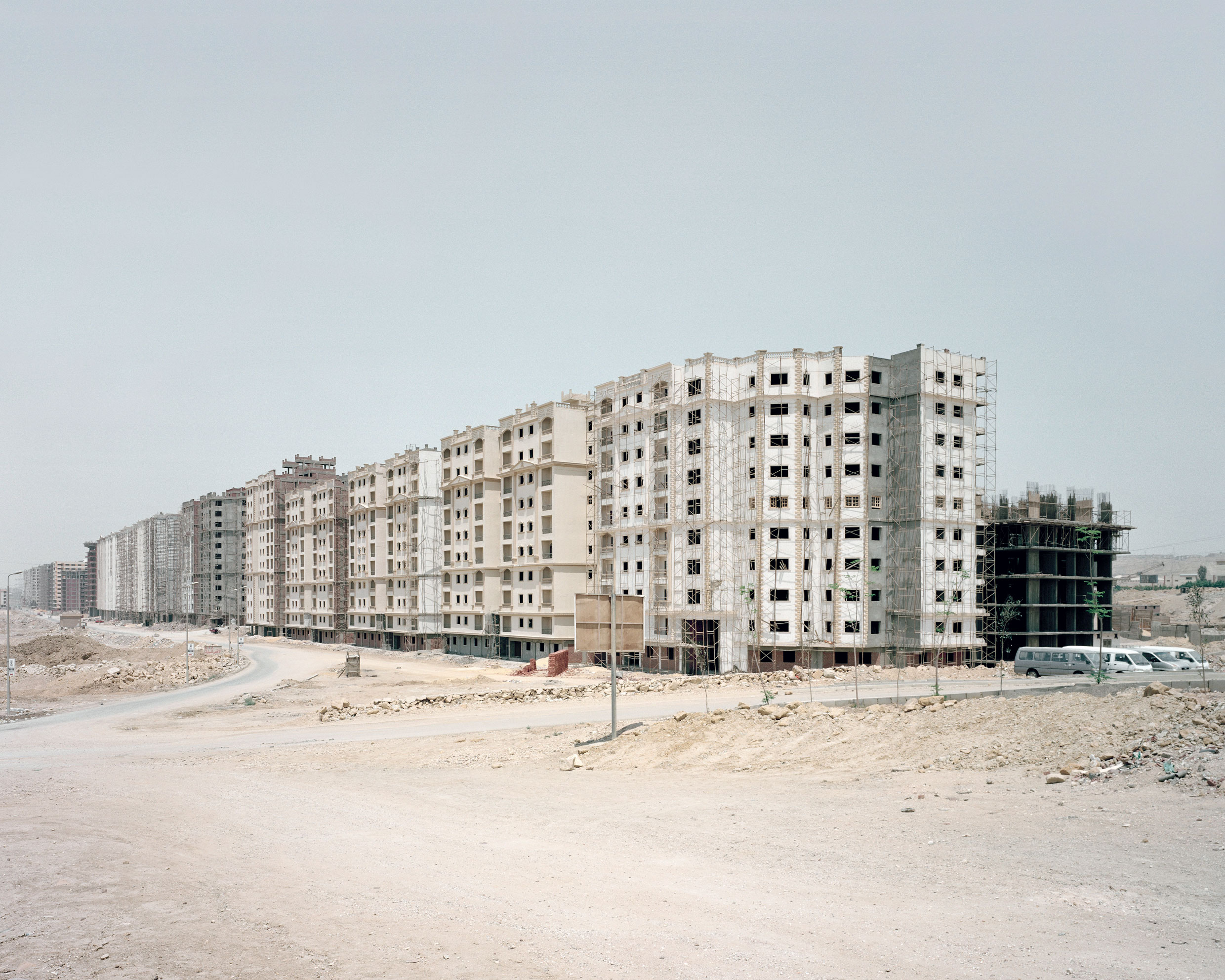 Cairo Ring Road, Ägypten 2015 / Anthony Hamboussi An der Ring Road von Kairo sind periphere Wohnungswüsten entstanden. Die Fotografien zeigen die exzessiven Ausmasse und desillusionierenden Auswirkungen auf die urbane Landschaft. Millionen von Quadratmetern mit leerstehenden Immobilien kreisen die Agglomeration von Kairo ein. Der boomende Wohnungsbau hat in den letzten Jahren zu einem Überangebot von überteuerten, aber dürftig konstruierten Wohnungen geführt. Für die lokale Bevölkerung sind sie geog