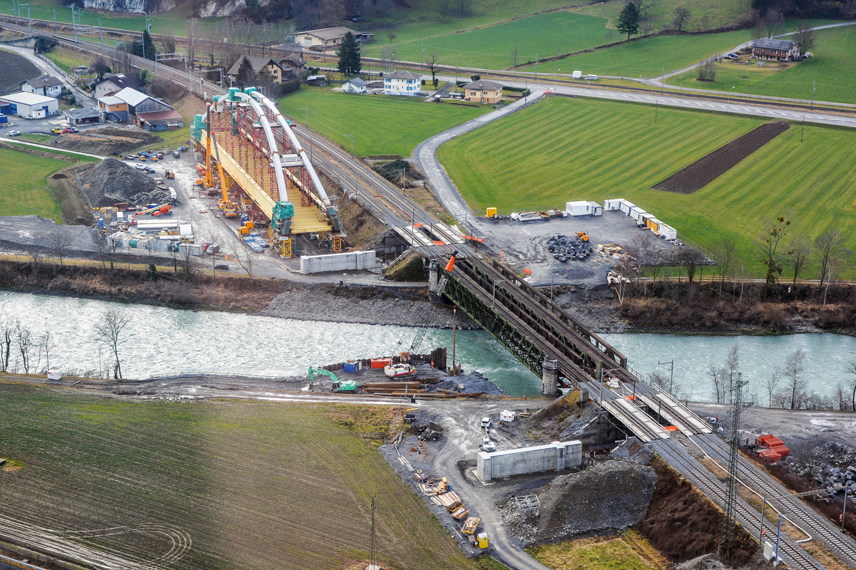 Stahlkonstruktion der neuen Brücke auf dem Installationsplatz mit den beiden Vorschublagern beidseits der Rhone und der Plattform für die provisorischen Pfeiler am rechten Ufer. Die beiden definitiven Widerlager entstehen in den Baugruben im Eisenbahndamm unter den vier Behelfsbrücken (Fliessrichtung von links nach rechts).