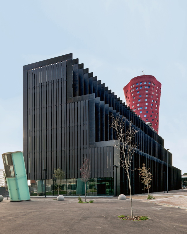 Das abgestufte Volumen des Neubaus war im Masterplan vorgegeben, RCR Arquitectes und Blázquez Guanter haben die Fassaden mit Stahlschwertern – dem eigentlichen Haupttragwerk – rhythmisiert. Das dahinter aufragende rote Hochhaus wurde vom japanischen Architekten Toyo Ito entworfen und dient als Hotel.