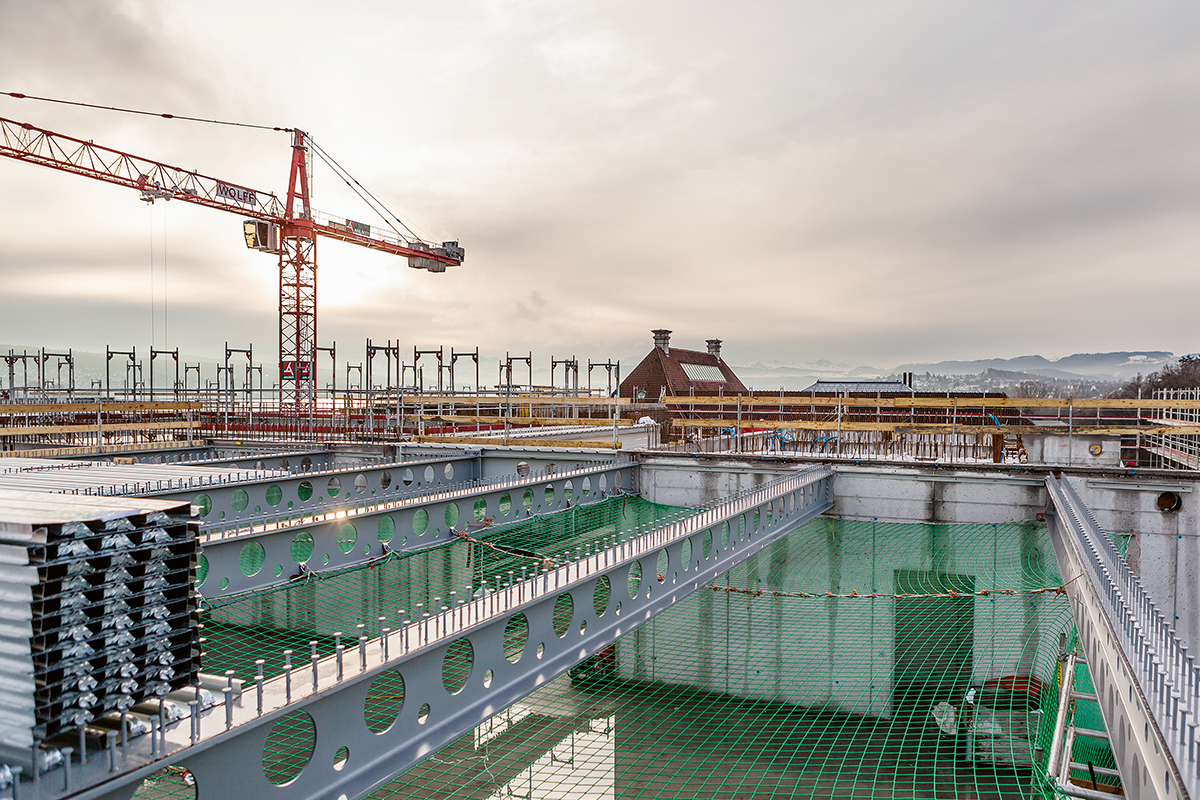 Perforierte Träger mit Kopfbolzen für den Verbund: Das Tragwerk des Gebäudes besteht über Terrain aus einem Skelettbau mit vorfabrizierten Stahlstützen und gedrungenen Stahl-Beton-Verbunddecken. Um die Medien in der reduzierten Konstruktionshöhe unterbringen zu können – die Konstruktionshöhe inklusive untergehängter Decke beträgt bei Spannweiten von bis zu 14 m nur 75 cm –, sind alle Träger systematisch perforiert.