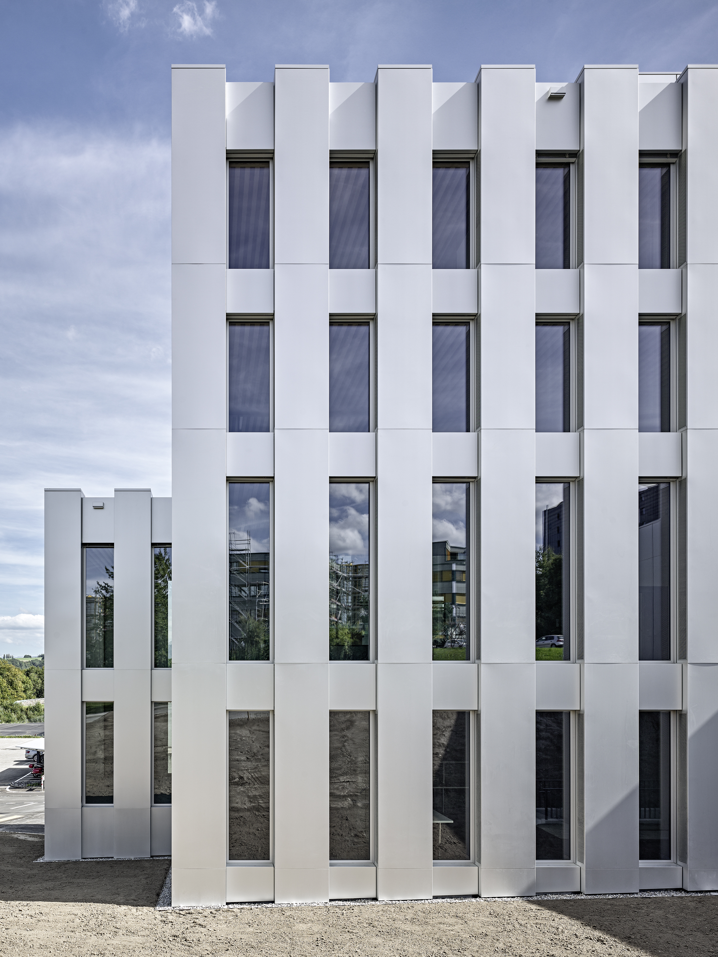 Premier Prix, Région Ouest: Bâtiment de la police cantonale fribourgeoise, Granges-Paccot FR, 2017 En raison du développement de la police cantonale fribourgeoise, le canton a planifié un ouvrage complémentaire sur une parcelle adjacente aux deux bâtiments existants. Ce nouveau volume comprend des bureaux, une cafétéria, des salles de conférence et d’entrainement, des locaux de stockage ainsi qu’un garage souterrain. Le revêtement sobre en aluminium intègre la façade à son environnement et