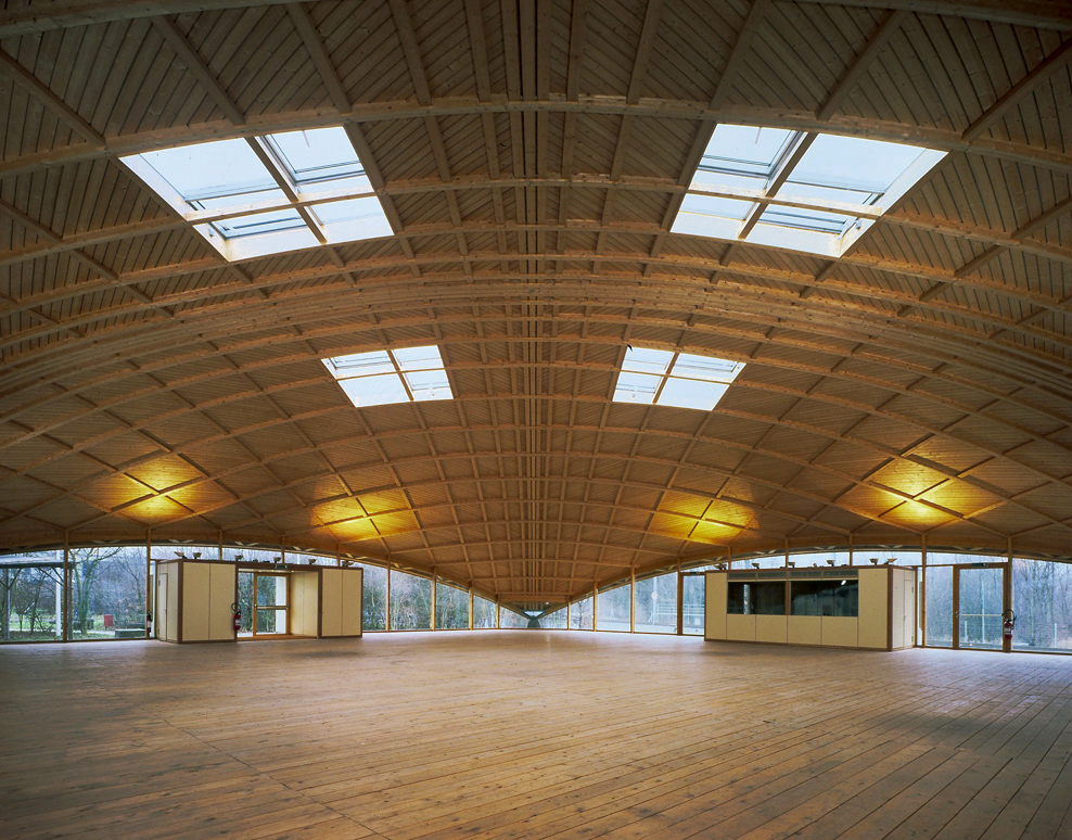 «Polydôme» in Brettstapelholz-Bauweise des Holzbauers Julius Natterer, Lausanne (1991). Die Holzrippenschale überspannt einen Grundriss von 25×25m mit einer Firsthöhe von 6.8m. Die Rippen bestehen aus vier Lagen Brettern 27/120mm, von denen in den Knotenp